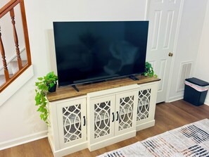 A television on a wooden cabinet with decorative glass doors, a potted plant, and a trash bin. - Walk to Shoppes of Kildaire & Close to Bond Park! (Cary)