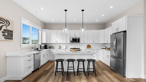 A modern kitchen with white cabinets, a stainless steel refrigerator, and a wooden floor. - Oceanview Retreat - Hot Tub + Game Garage + Spa Suite + Kids Cave - Westport, WA (Westport)