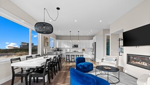 A modern kitchen with white cabinets, a dining area with a large table and chairs, a living room with a fireplace, and a large window overlooking a landscape. - Oceanview Retreat - Hot Tub + Game Garage + Spa Suite + Kids Cave - Westport, WA (Westport)