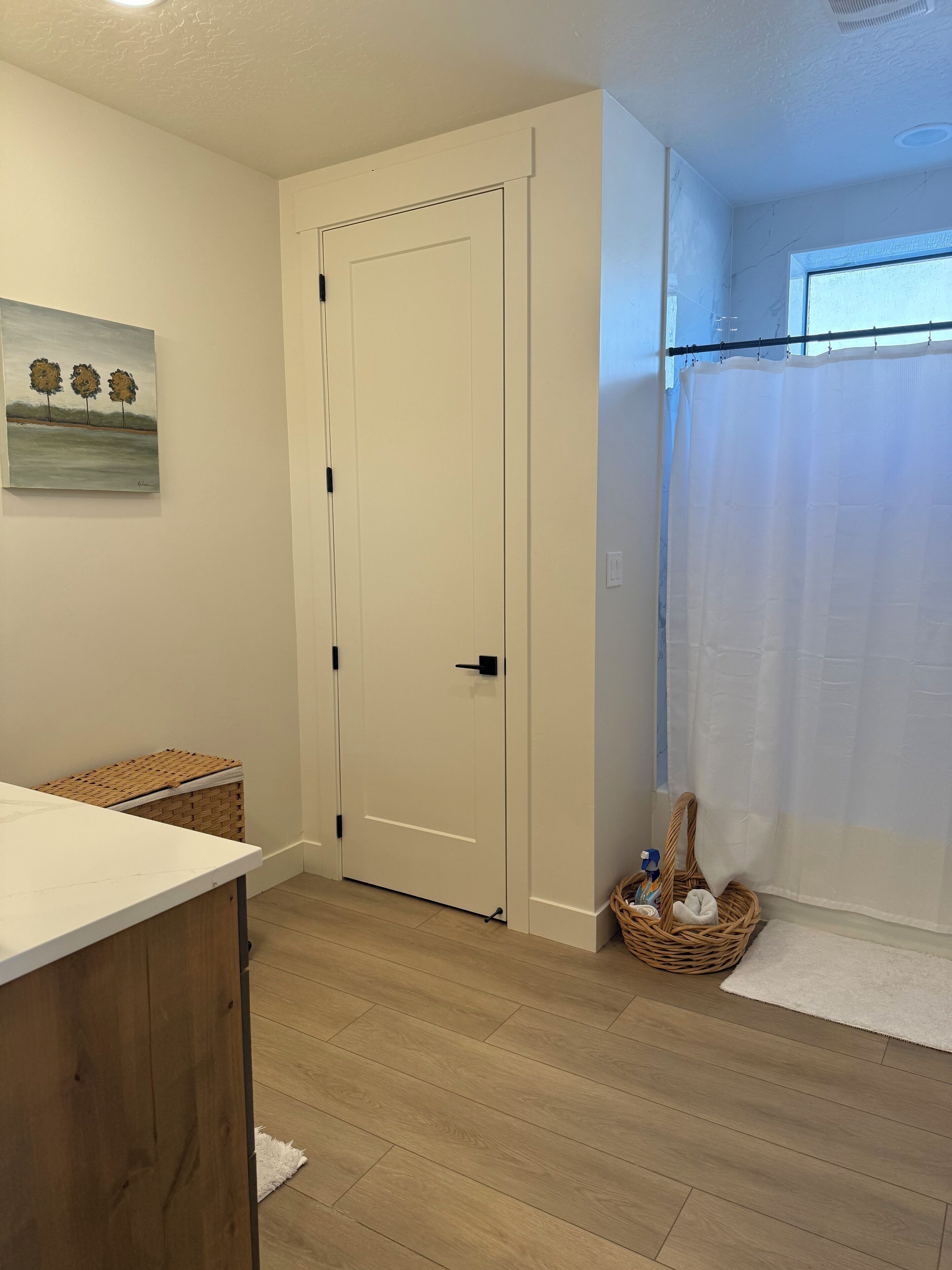 Une pièce avec un plancher en bois, une porte blanche, un mur blanc orné d’un tableau, un panier en osier et un rideau blanc.