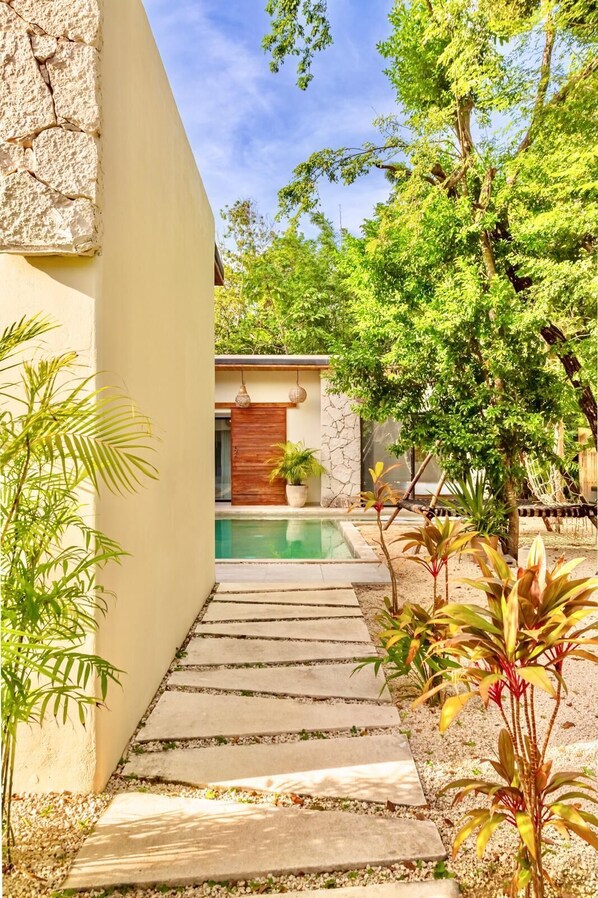 Un sendero conduce a una zona de piscina con un muro de piedra, plantas en macetas y una puerta de madera.