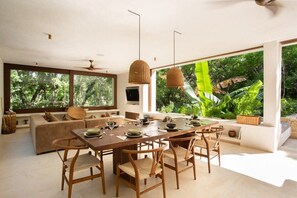 A modern living room with a large dining table, comfortable seating, and large windows offering a view of lush greenery. - Tepui: Natural Luxury with Pool & Rooftop (Tulum)