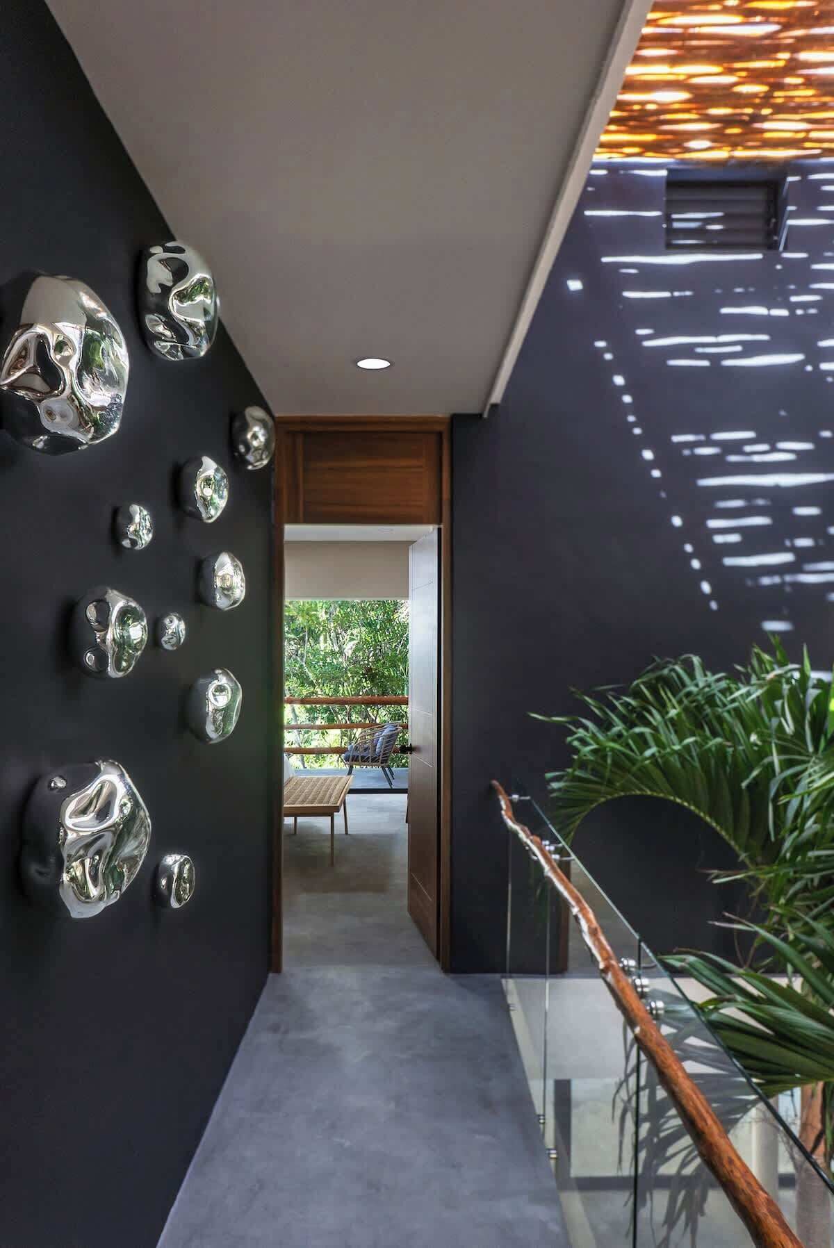 Un interno moderno con un corridoio che conduce a una zona pranzo, caratterizzato da una parete con oggetti sferici riflettenti, una ringhiera in vetro e vista su piante verdi all'esterno.