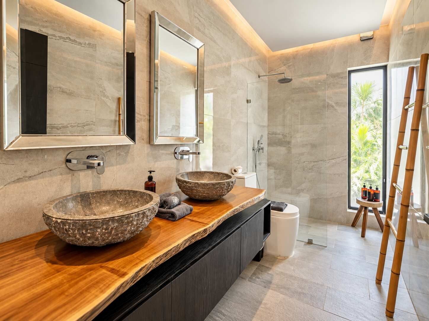 A modern bathroom with two large stone bowls on the vanity, a walk-in shower, and a view of greenery through the window.
