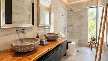 A modern bathroom with two large stone bowls on the vanity, a walk-in shower, and a view of greenery through the window.