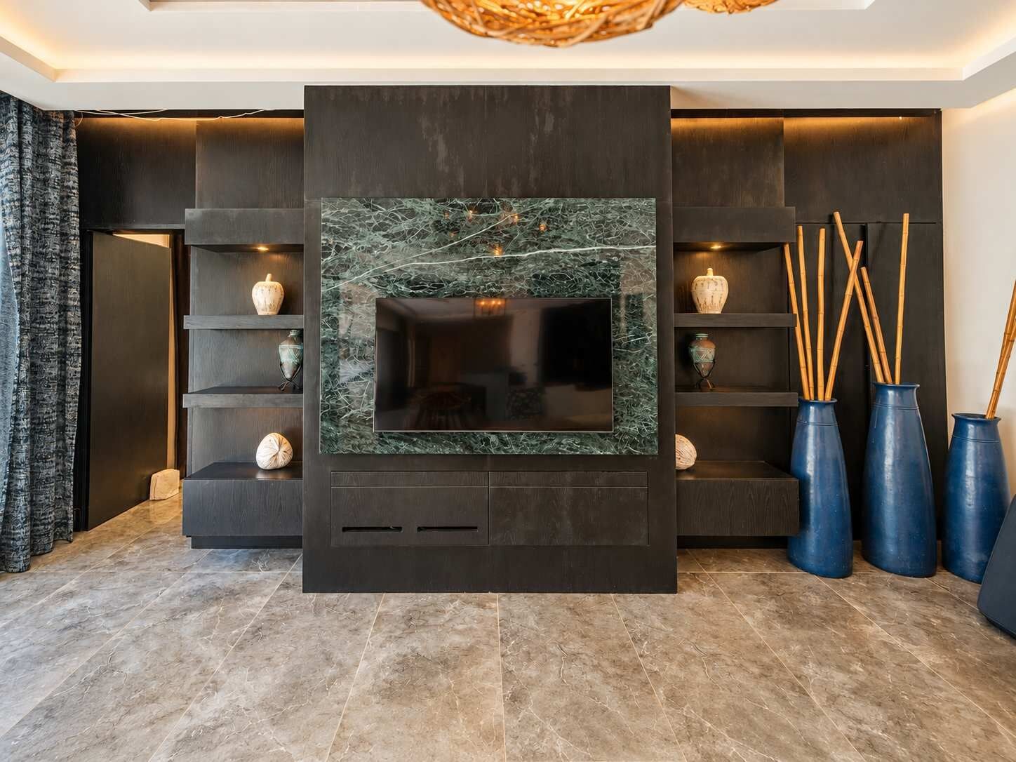 A modern living room with a large flat-screen TV mounted on a dark wall with a marble-like texture. There are shelves with decorative items and vases on the right side.