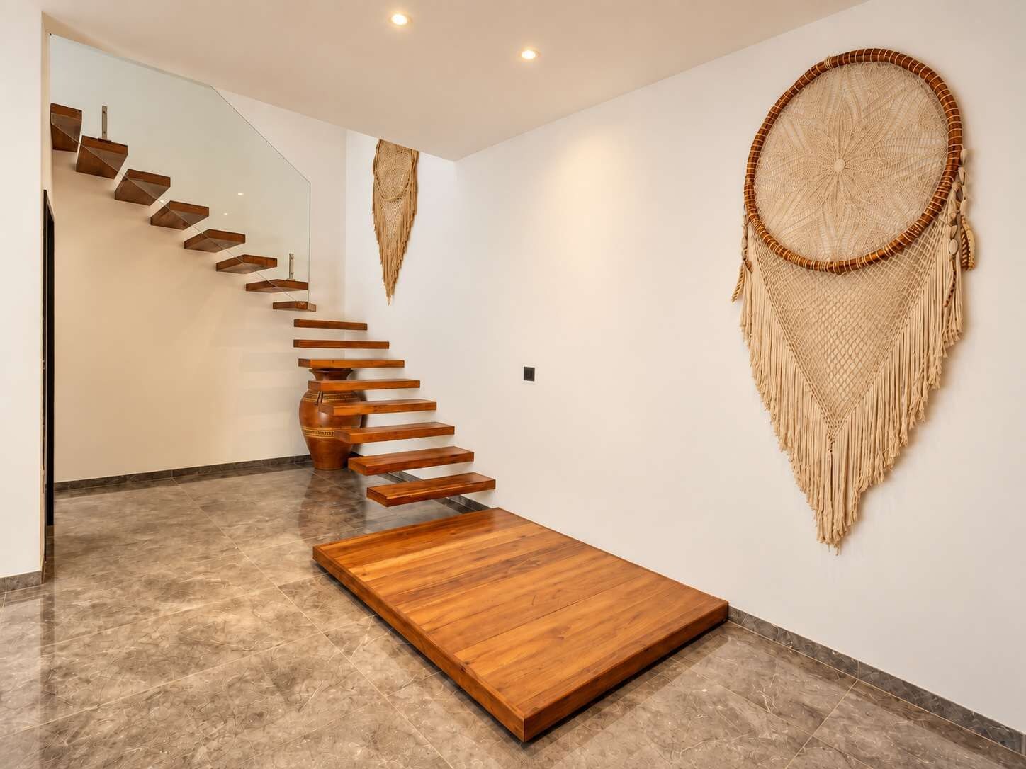 Un interno moderno con una scala in legno che conduce ad una piattaforma, un grande dreamcatcher intrecciato appeso al muro e un pavimento in marmo.