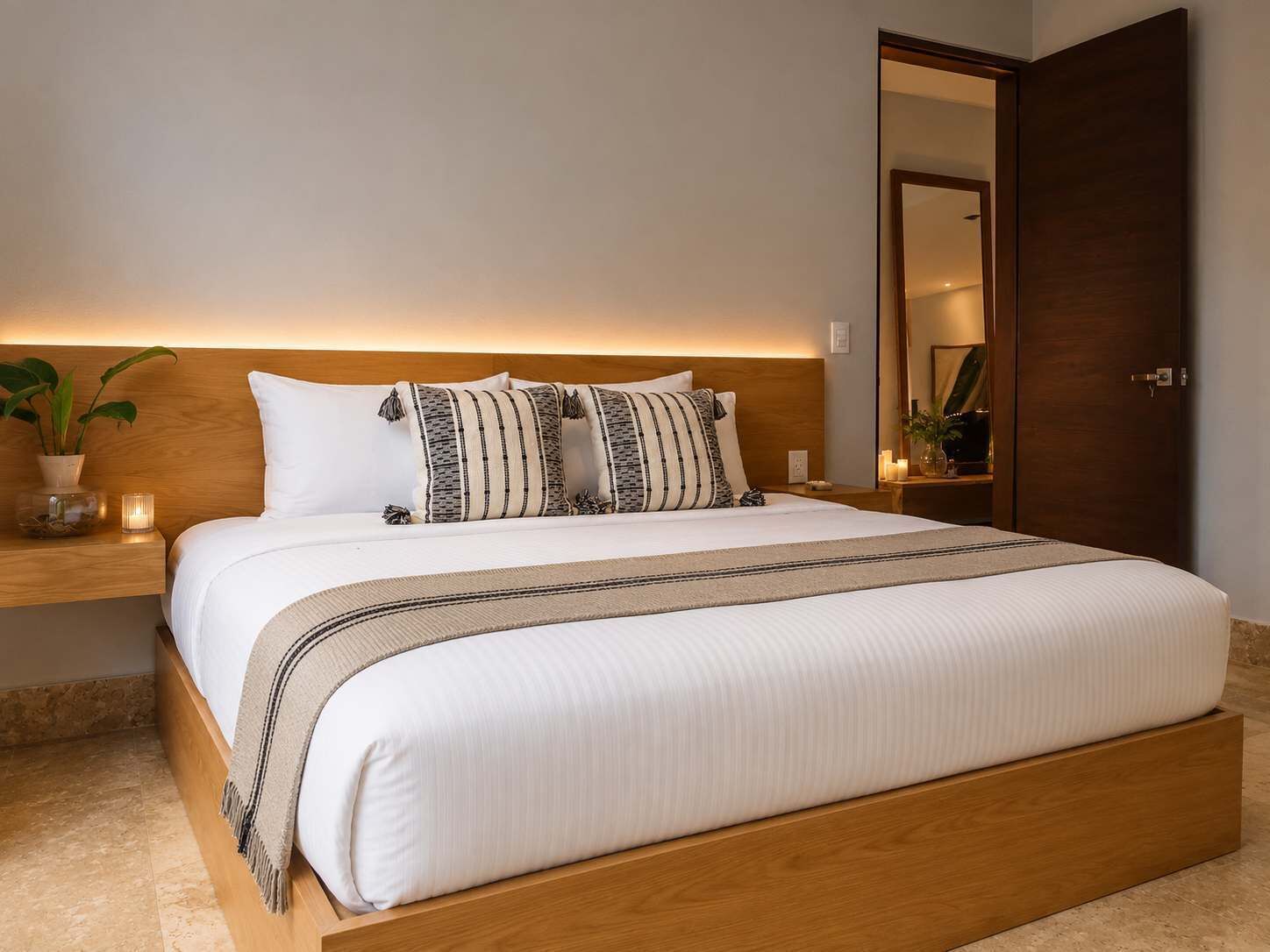 Ein ordentlich bezogenes Bett mit Holz-Kopfteil, ein Nachttisch mit Pflanze und ein Spiegel an der Wand.