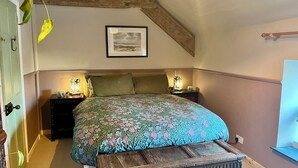 Ein Schlafzimmer mit einem Bett, zwei Nachttischen, einem kleinen Tischchen, einem gerahmten Bild an der Wand und einer Holzbalkendecke.