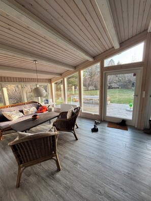 Une pièce spacieuse avec un plafond en bois, de grandes fenêtres, un canapé, deux fauteuils en osier, une table et une porte donnant sur l’extérieur.