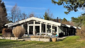A modern house with a large porch, surrounded by a grassy yard and trees.