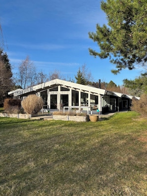 A modern house with a large porch, surrounded by a grassy yard and trees.
