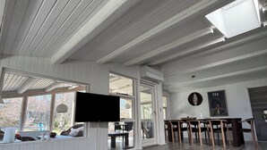 A spacious room with a high ceiling, large windows, a round wooden table, and a flat-screen TV mounted on the wall.