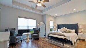 Une chambre moderne avec un lit, deux fauteuils, une petite table, un ventilateur de plafond et de grandes fenêtres.
