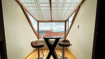 Um quarto pequeno com uma mesa e dois banquinhos, uma janela com vista para um edifício com cúpula, e um teto com tecido translúcido.