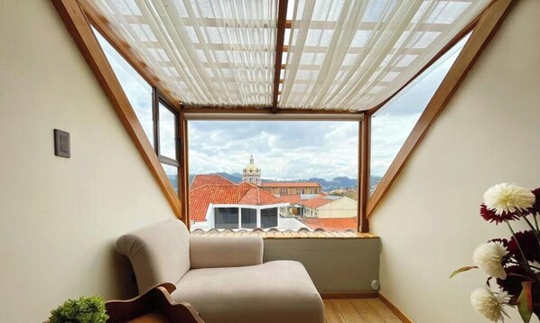 A room with a sloped ceiling, a window with a view of buildings, a sofa, and a potted plant. - Hotel Ensueños by HHG (Cuenca)