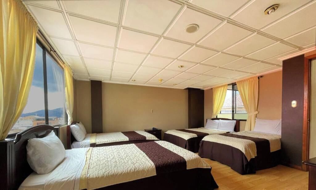 A hotel room with three beds, a large window with curtains, a wooden floor, and a ceiling with recessed lighting.
