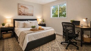 Un dormitorio con cama, mesitas de noche, escritorio, silla, ventana y un cuadro en la pared.