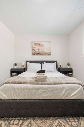 Een keurig opgemaakt bed met een bedsprei met patroon, twee nachtkastjes met lampen, een aan de muur hangend schilderij, en een kleine pot met een plant aan weerszijden van het bed.