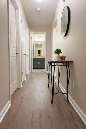 A hallway with white doors, a wooden floor, a small table with a plant, and a mirror on the wall. - The Rally Point @ Antietam (Columbus)