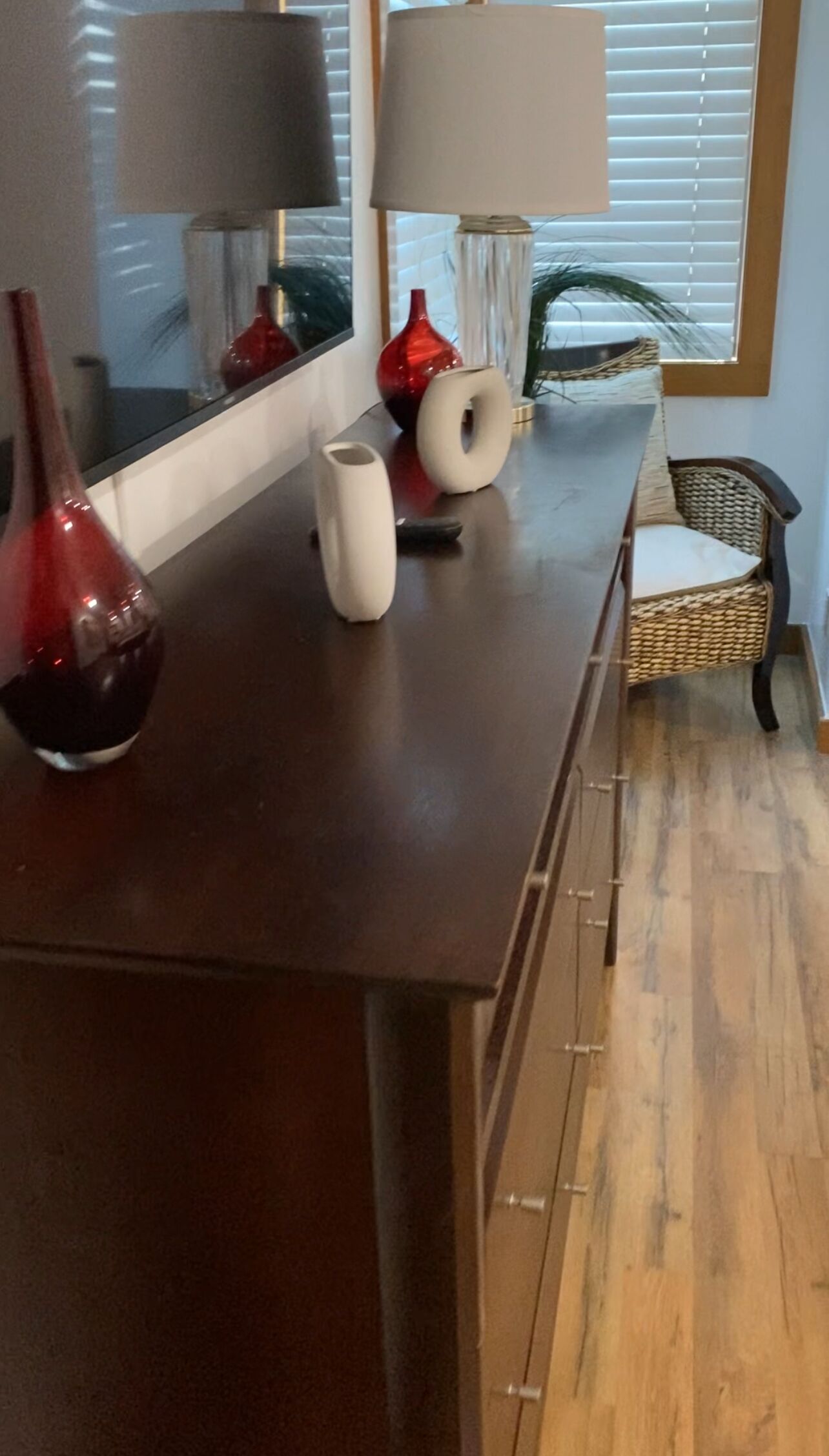A dark wooden dining table with a vase, a white pitcher, and a red vase. There is a wicker chair and a window with blinds in the background.