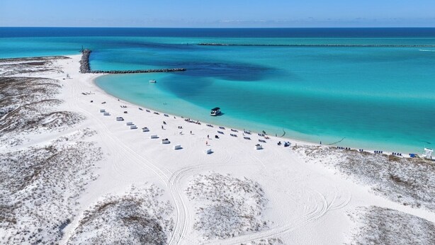 Vista aérea de una playa con un mar turquesa cristalino, una orilla arenosa y un muelle que se adentra en el agua.