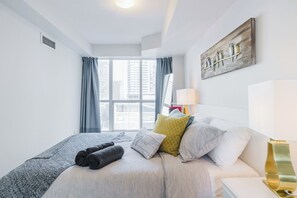 Ein modernes Hotelzimmer mit einem großen Bett, einem Fenster mit Vorhängen, einem Wandbild und einer Nachttischlampe.