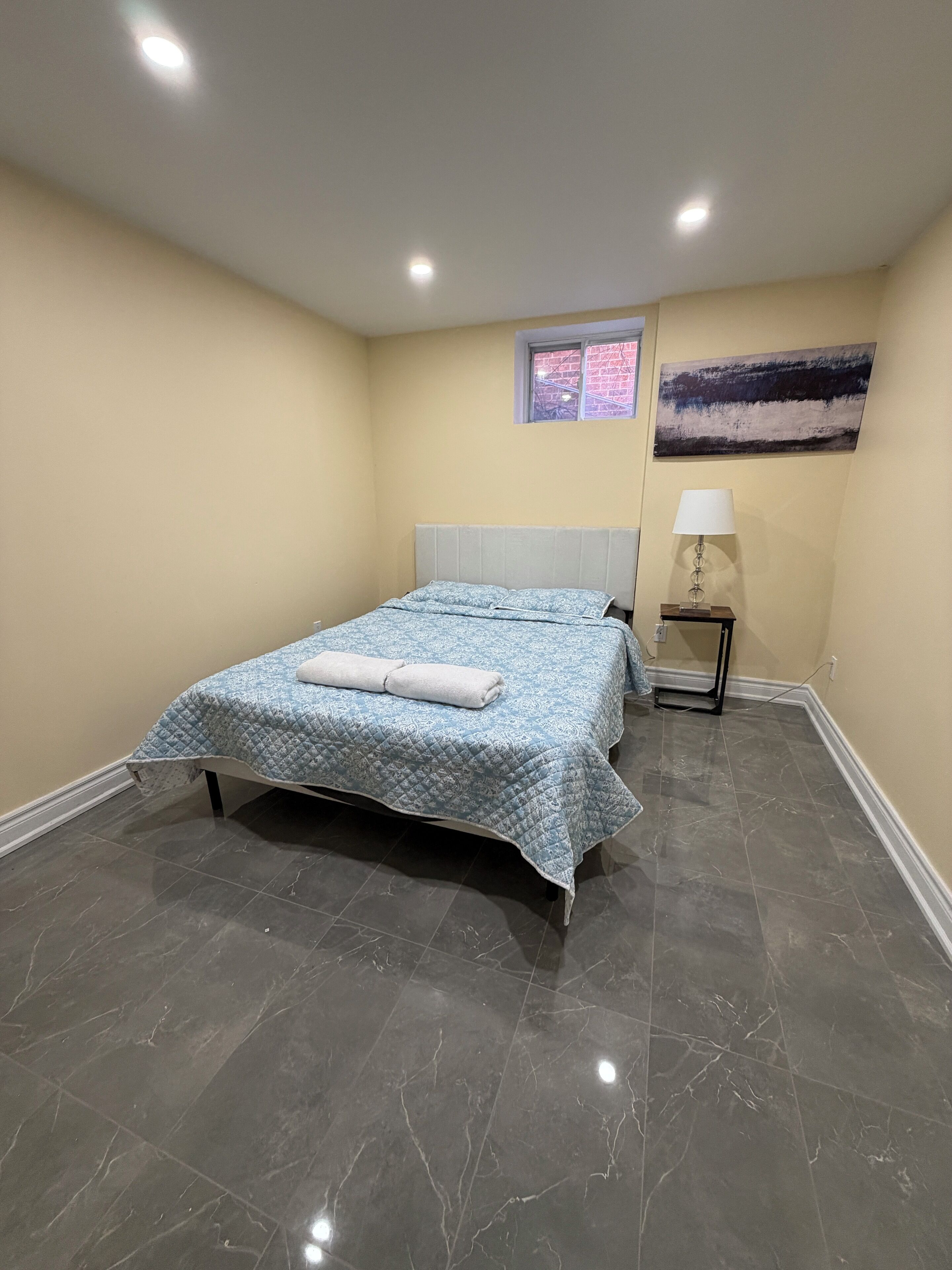 A bedroom with a bed, a nightstand, a lamp, a window, and a painting on the wall.
