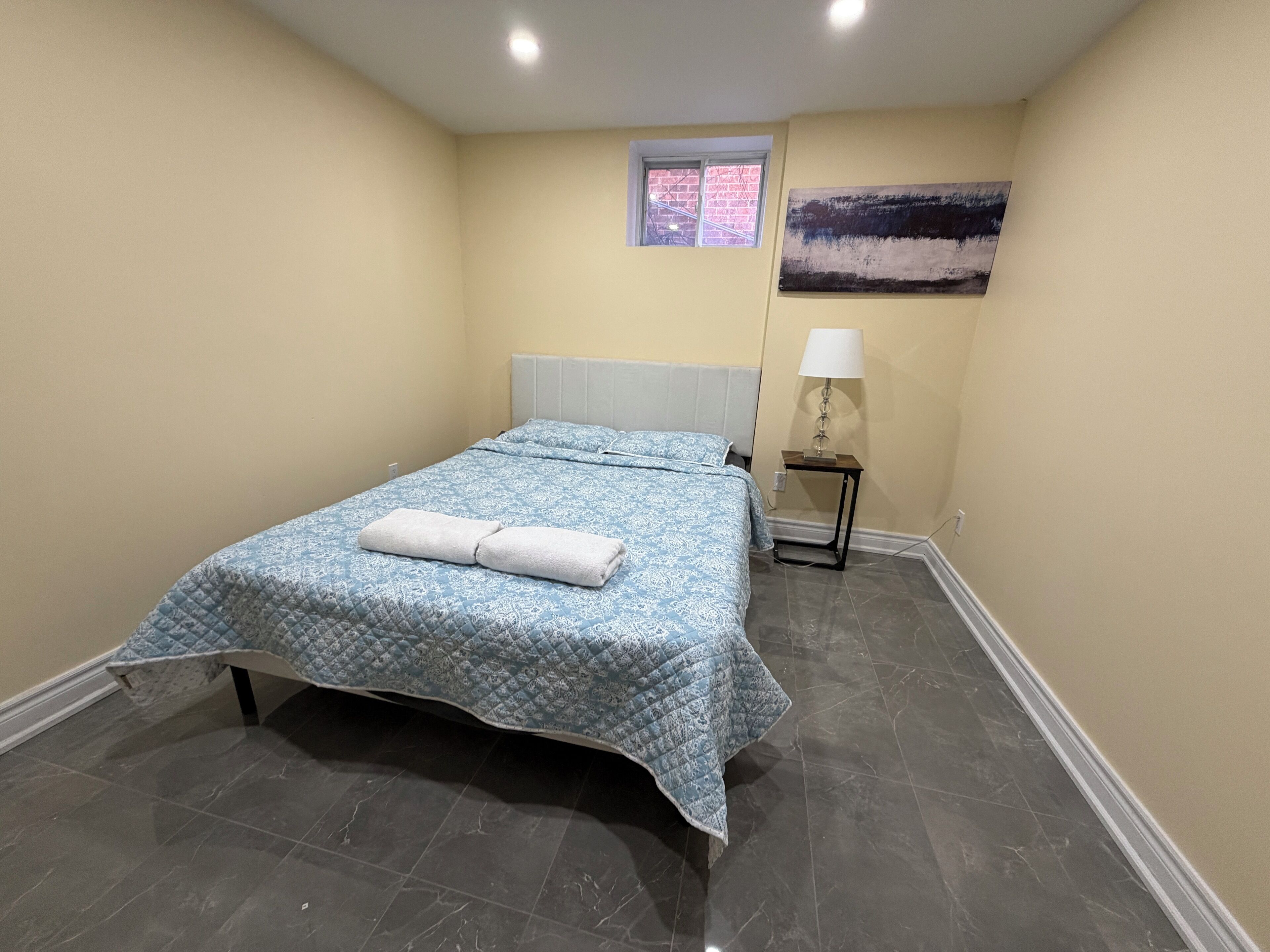 Una camera da letto con un letto, un comodino, una lampada, una finestra e un quadro appeso al muro.