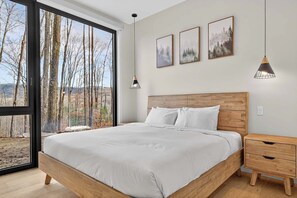 A bedroom with a large window, a wooden bed, a nightstand, and three framed pictures on the wall. - NEW! Forêt de Verre | 7 Min to Village | Hot Tub, Firepit & Entertainment | 16 Guests (Mont-Tremblant)