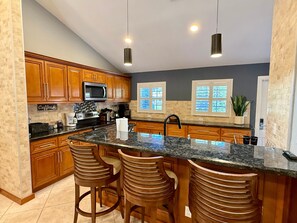 A kitchen with wooden cabinets, a granite countertop, a stainless steel microwave, a coffee station, and a window with blinds.