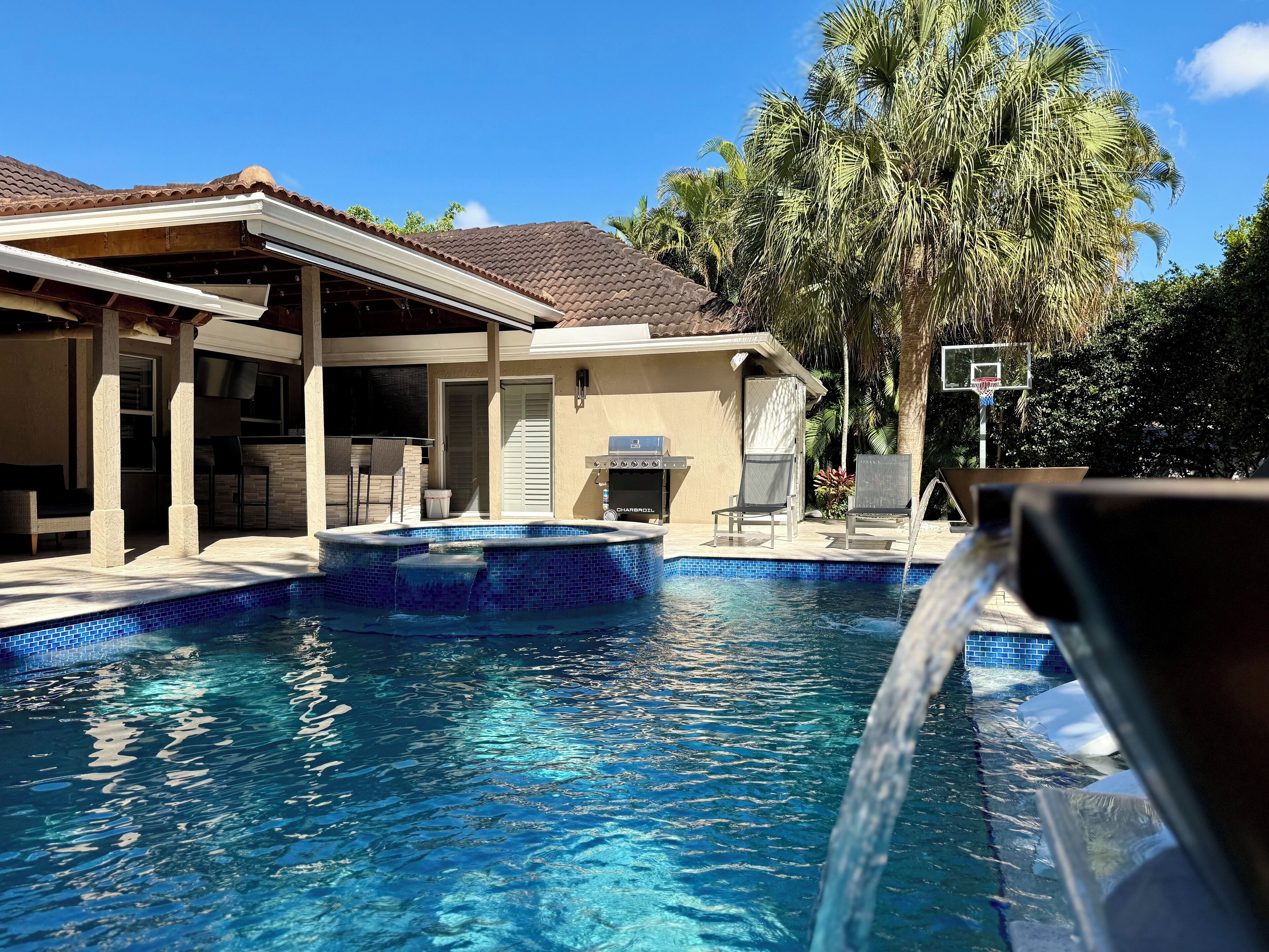 A swimming pool with a tiled deck, a covered patio area, and a grill.