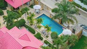 Una piscina con sillones de descanso y sombrillas, rodeada de palmeras y un edificio de techo rojo.