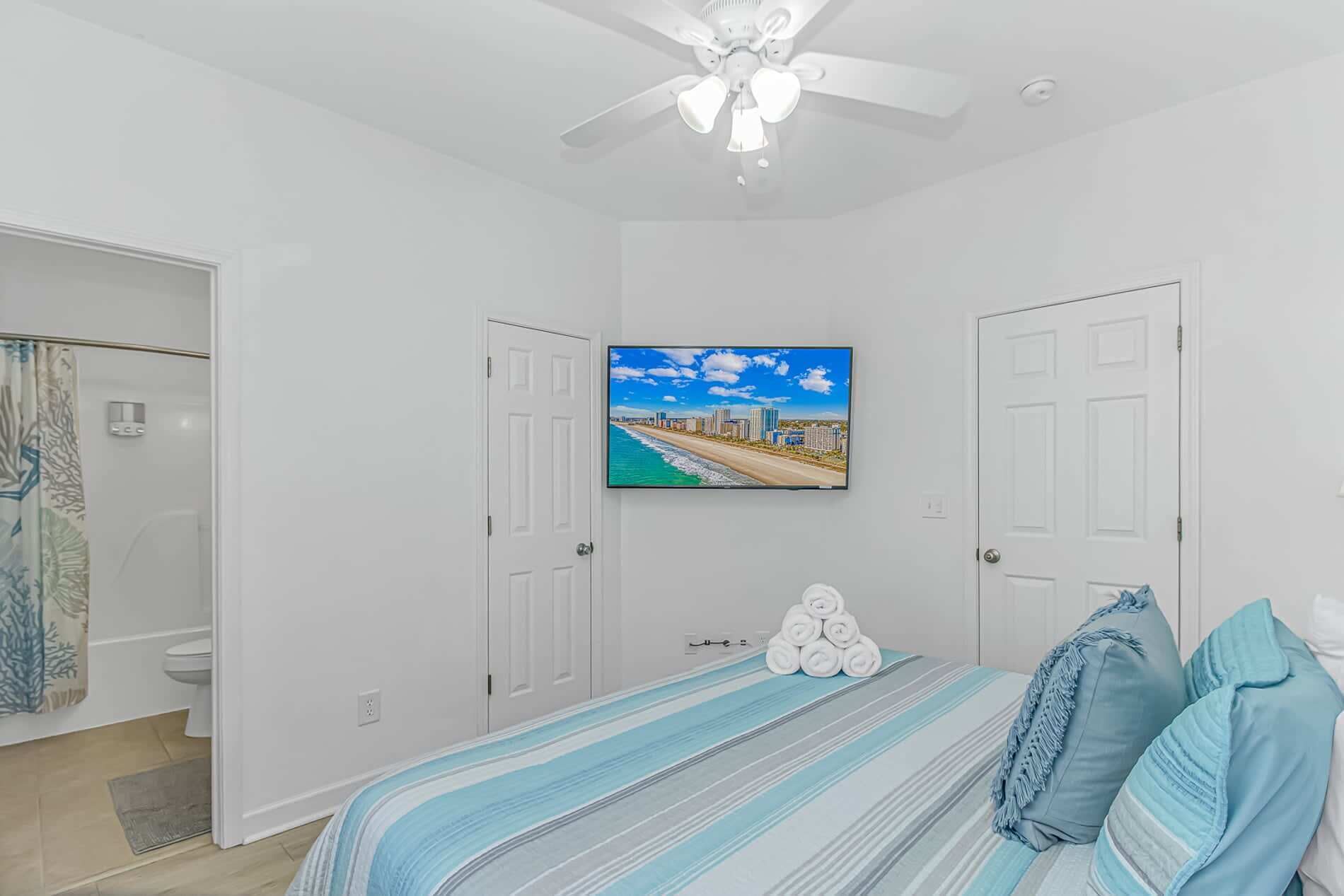 A bedroom with a bed, a ceiling fan, a TV on the wall, and a door leading to a bathroom.