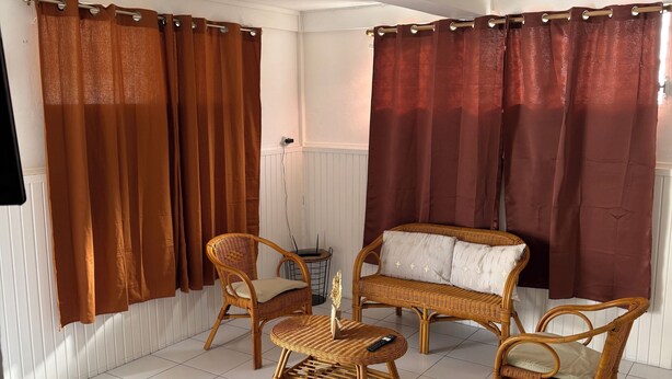 A room with a white tiled floor, brown curtains, wicker furniture, and a small table with a decorative item. - Residence with sea view and close to beaches (Le Marin)