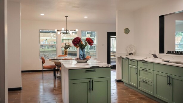A modern kitchen with green cabinets, a central island with a vase of flowers, and a dining area with chairs. - Private Pool • Movie Theater • Sleeps 12 / 4BR (San Antonio)