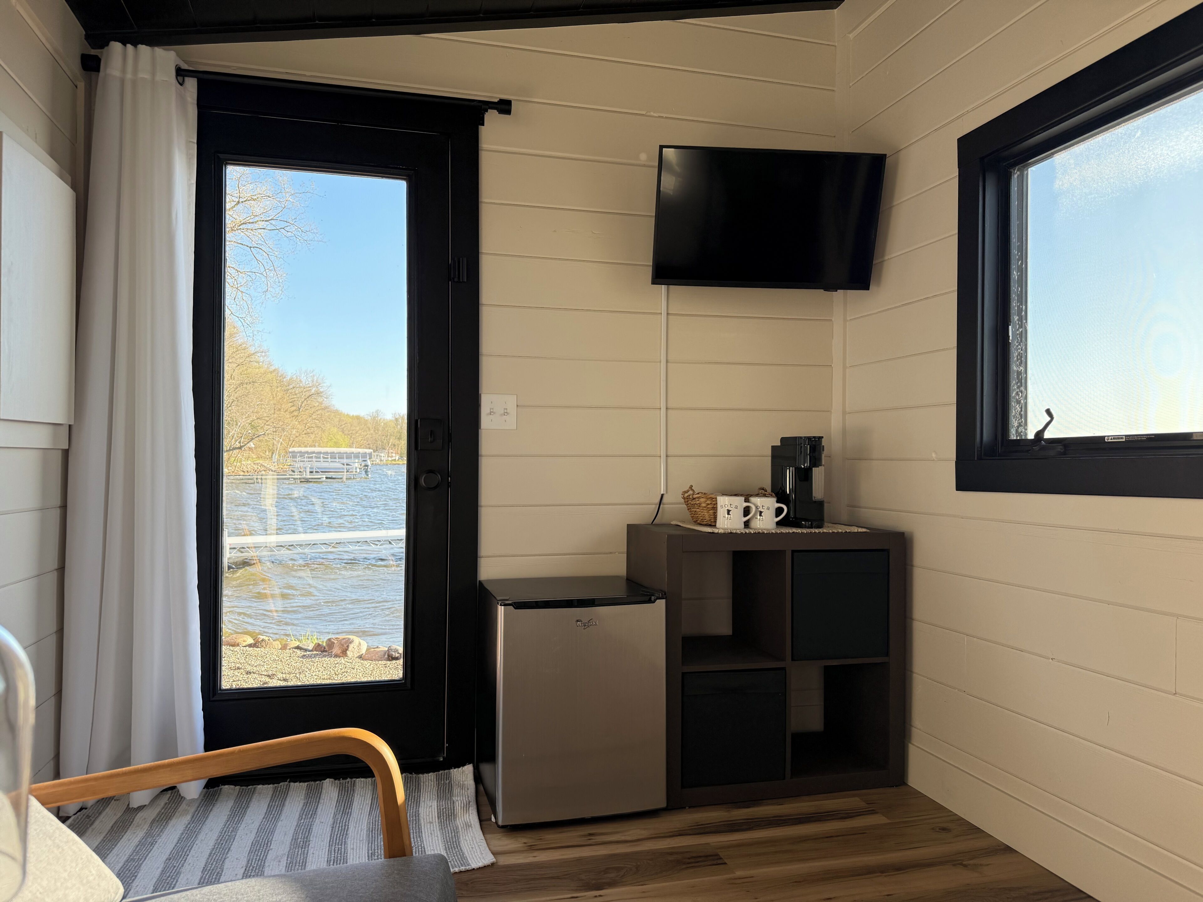 A compact room with a door leading outside, a small refrigerator, a flat-screen TV mounted on the wall, and a wooden chair.