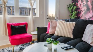 Ein modernes Wohnzimmer mit einem roten Sessel, einem schwarzen Sofa mit vielen Kissen, einem runden Couchtisch mit Pflanze und Fernbedienungen sowie einem großen Fenster mit Vorhängen.