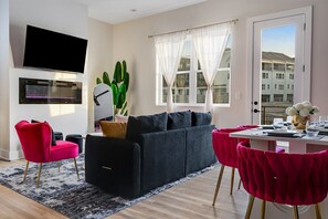 A modern living room with a black sofa, red chairs, a flat-screen TV, a fireplace, and a large window with sheer curtains. - NEW!Pink Palace:Rooftop Hot Tub, Pool, Arcade, Beer Pong, Karaoke,BBQ, Game Room (nashville)
