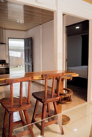 A wooden bar stool set with a wooden table, a kitchen area with a stove, and a doorway leading to a bedroom. - La maison Minou (Palmar Norte)