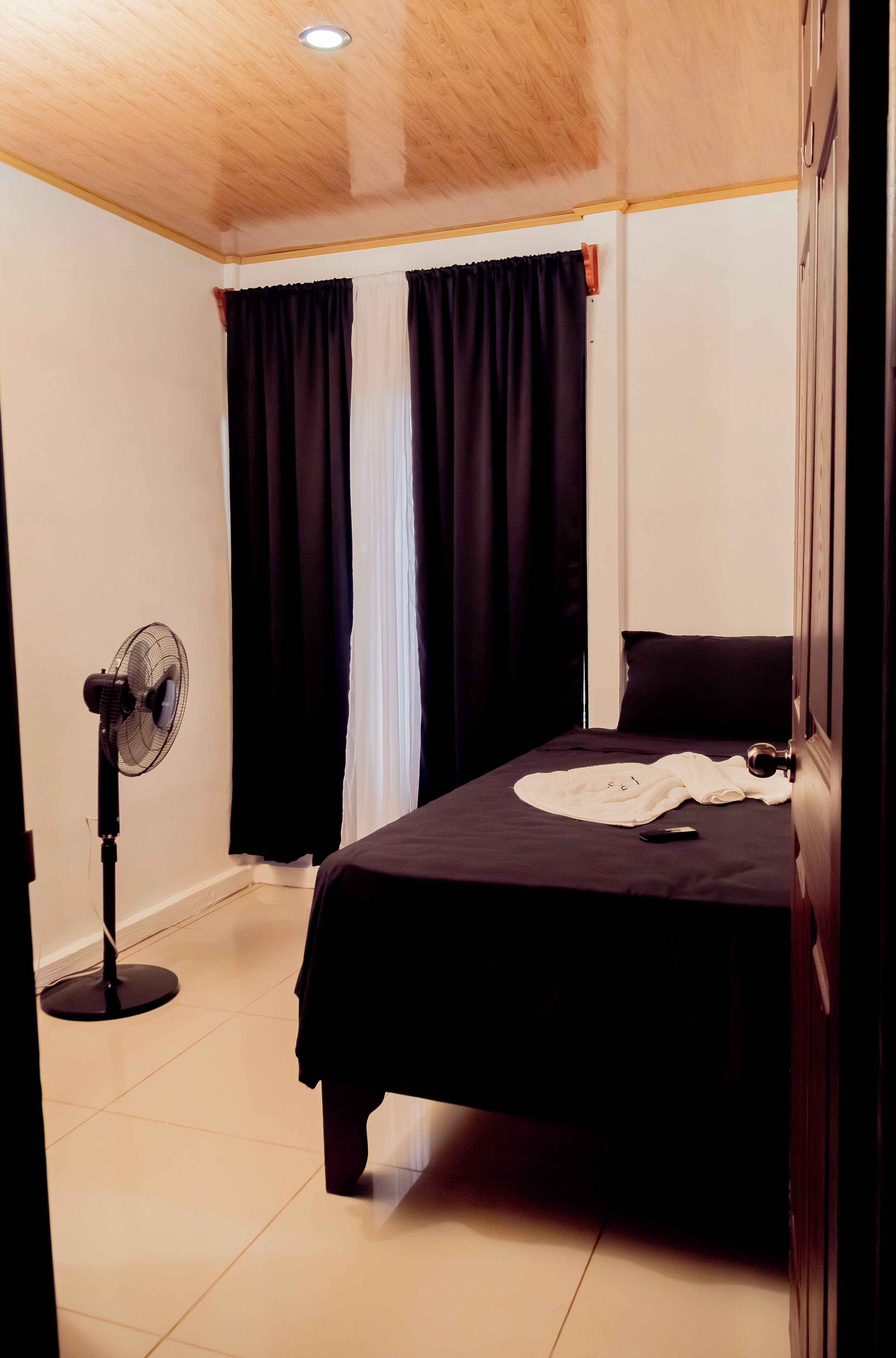A bedroom with a bed, a standing fan, and a window with curtains.