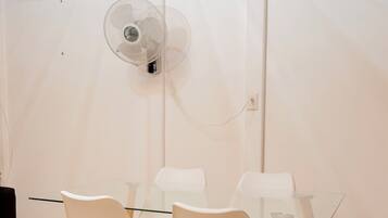 Una habitación con una mesa de vidrio, sillas blancas, un ventilador de pie y un equipo de aire acondicionado en la pared.
