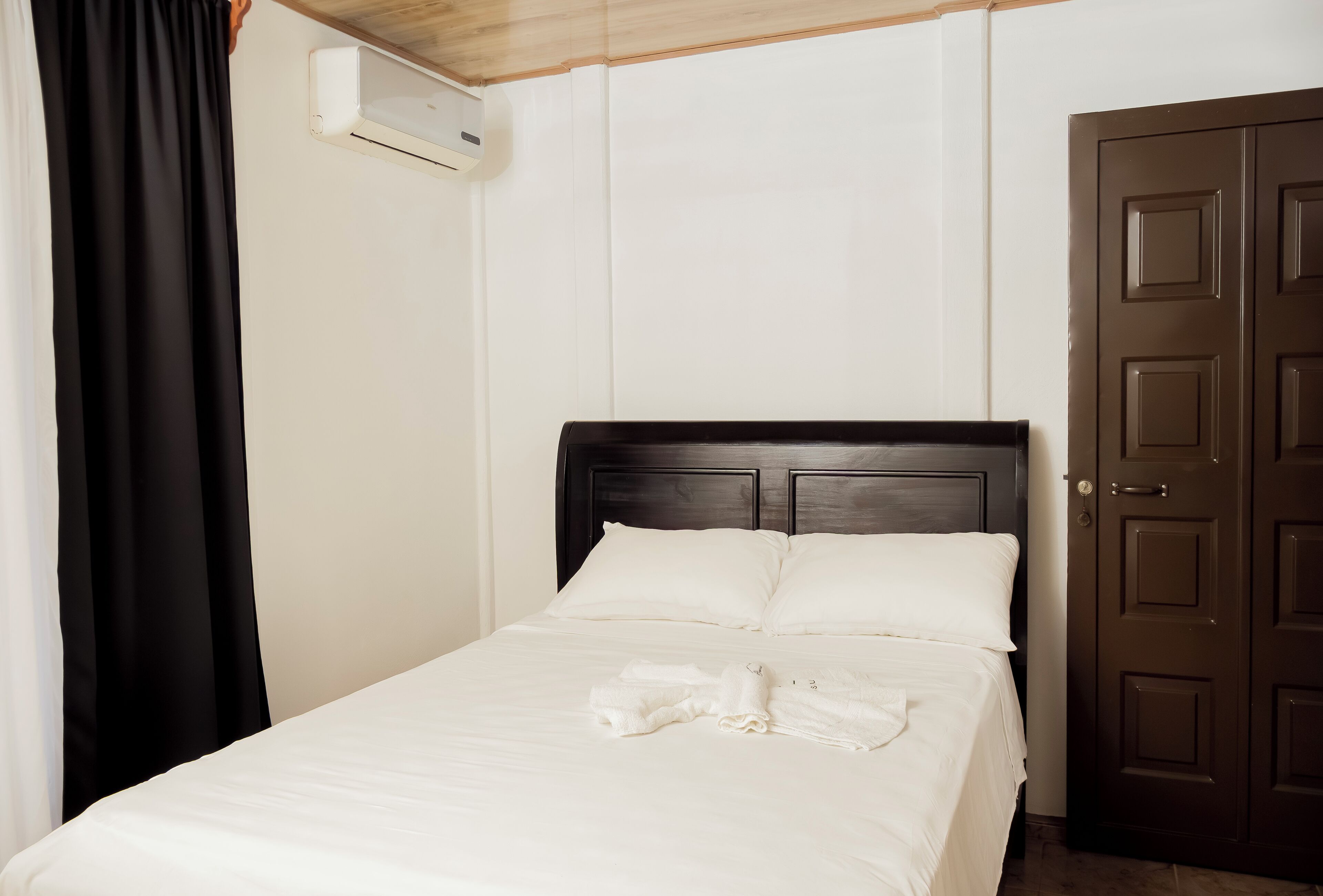 Un lit bien fait, avec du linge de lit blanc et une serviette pliée, une tête de lit en bois, une porte marron foncé fermée et un appareil de climatisation fixé au mur.