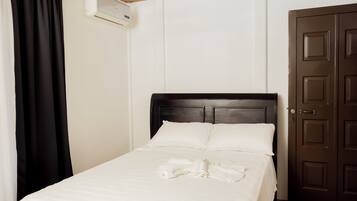 Un lit bien fait, avec du linge de lit blanc et une serviette pliée, une tête de lit en bois, une porte marron foncé fermée et un appareil de climatisation fixé au mur.