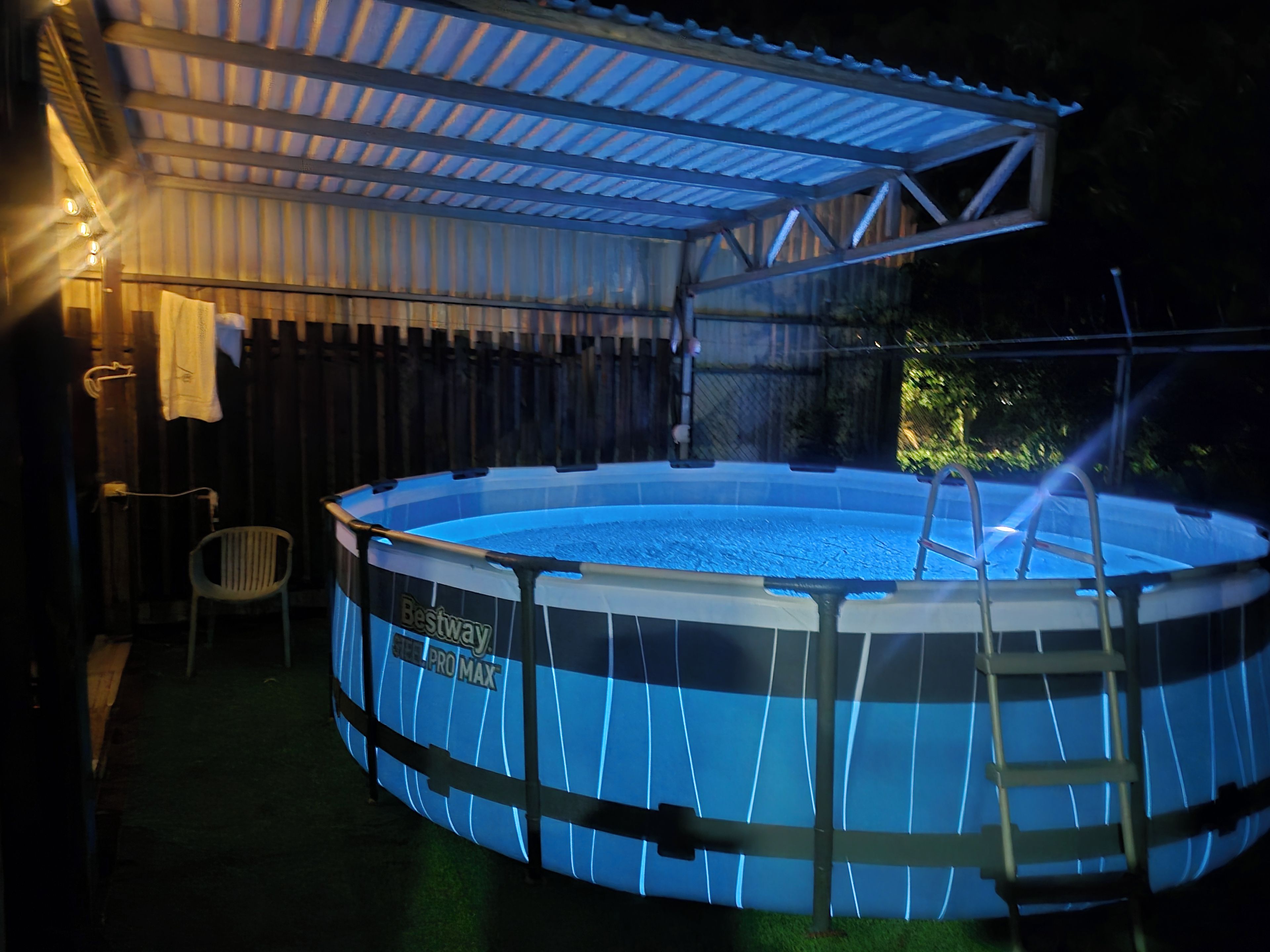 An outdoor swimming pool with a ladder, surrounded by a covered area with a roof.