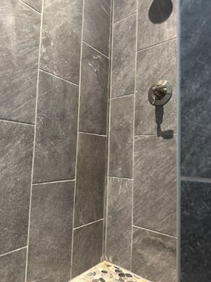 A corner of a room with dark grey tiled walls and a shower area with a visible showerhead.