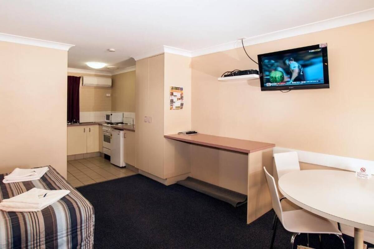 A compact hotel room with a kitchenette, a dining area, a TV mounted on the wall, and a bed with a striped blanket.