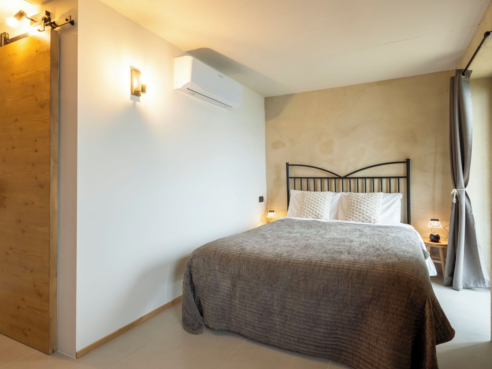 A bedroom with a bed, a wall-mounted air conditioner, a bedside lamp, and a wooden door.