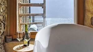 Una habitación con cama, ventana con vista a la ciudad y montañas, una botella de vino y dos copas sobre una mesita.