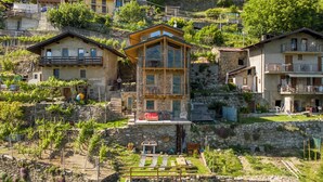Un pueblo en una colina con casas de piedra, un edificio central con balcón y un viñedo.